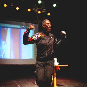 Courage pumps her arms up while stepping sideways. She is a middle aged black woman with closed croped curly hair.She is wearing a black tracksuit and white runners. A video screen is behind her on stage.