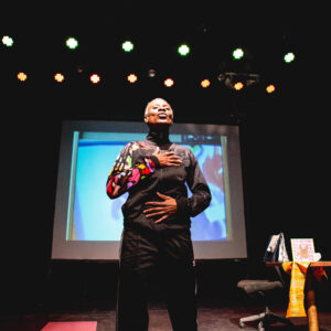 Couragehas both hands on her diaphram with her lips pursed in a breathing excercise. She is a middle aged black woman with closed croped curly hair.She is wearing a black tracksuit and white runners. A video screen is behind her on stage.
