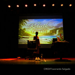 On blacked out stage: Courage is wearing a white dress standing behind a black chair. She is backlit by a bright video screen behind her. Video image shows a tropical scene with words "Rebirth of Courage".